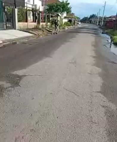 The video shows a wide-angle view of a residential street in Roza, Bulgaria. The street is paved with asphalt, and there are visible cracks and patches of moisture, suggesting it might have recently rained or been wet. Puddles of water are also visible on the sides of the road.

On the left side of the street, a person dressed in dark clothing is operating a brush cutter or similar landscaping machine. They are moving away from the camera, clearing vegetation and debris along the curb and adjacent to fences and houses. The houses are mostly one or two stories, with fences separating them from the street.

Further down the street, more houses and trees are visible, extending into the distance. The sky appears clear, and the overall lighting suggests it is daytime. The scene depicts a routine maintenance activity in a quiet residential area. No text is visible in the video.