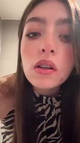 A young woman is applying lip liner while looking in a mirror. She is wearing a black and white print top and has long brown hair. She is holding a makeup pencil in her right hand and is lining her upper lip. The pencil says "Beauty Clever" on the side. She has a nose ring and braces. The background appears to be a bathroom.