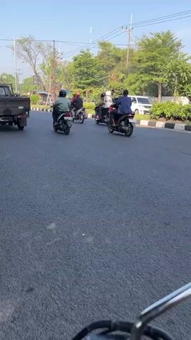 This media file captures a street scene in Klurak, Indonesia, likely during daytime given the bright sunlight and clear skies. The primary subjects are several individuals riding motorcycles on a paved road.

The scene depicts a road with traffic, including a black pickup truck in the foreground on the left, and a white van further down the road on the right. The motorcycles are moving in the same direction. One motorcycle in the foreground has two riders, with the driver wearing a helmet. Further ahead, other motorcycles with one or two riders are visible, all similarly equipped with helmets. The road is lined with trees and vegetation on both sides, suggesting a somewhat suburban or semi-urban environment. In the background, there are utility poles and wires, typical of a developed area.

The overall activity is transportation, with people commuting or traveling by motorcycle. The time of day appears to be morning or afternoon, judging by the quality of light and the shadows cast. The weather seems pleasant, with no indication of rain or overcast skies. There are no discernible emotions expressed as the focus is on the movement and the environment. No visible text is present in the frame. The location context of Klurak, Indonesia, places this scene within that specific geographical area.