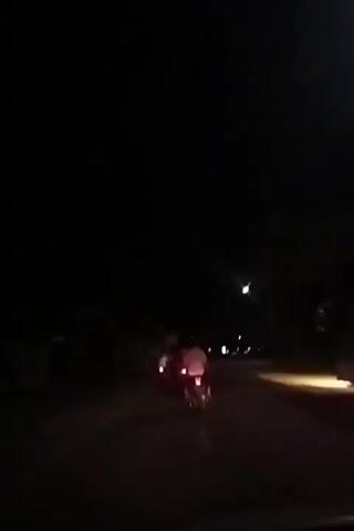 At night, a road with streetlights has a motorcycle on the left and a bus on the right. The bus is illuminated with multiple lights. The camera is positioned on or near the road surface, and the bus recedes into the distance.