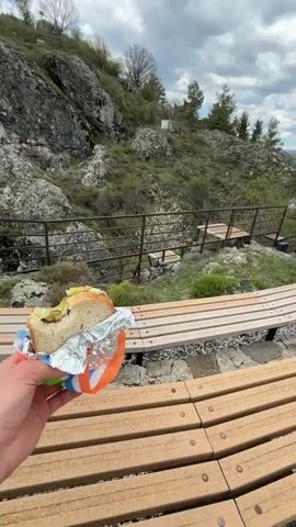 The video shows a person holding a partially eaten sandwich in their hand, sitting on a wooden bench. The camera pans up to reveal a rocky landscape with a metal railing, overlooking a green valley with rolling hills under a cloudy sky. The person is eating their sandwich while enjoying the scenic view from the elevated location.