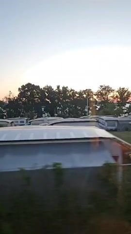 The video captures a view from a moving vehicle, possibly a train or car, looking out at a campground. The foreground is dominated by green grass, a blurry white object, and a yellow pole with some blue attached to it. In the mid-ground, several mobile homes or caravans are visible, parked on grassy plots. Some have awnings extended. Behind the mobile homes, a line of trees forms a backdrop, with a body of water and a hint of a sunset sky visible beyond them. The sky is a pale gradient of blue and white, suggesting either early morning or late evening. No people are clearly visible, and no specific actions or interactions are discernible due to the motion and distance. The overall impression is a peaceful, somewhat rustic scene at a campsite.