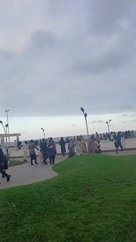The video shows a park area next to a beach on a cloudy day. Numerous people are walking along the paved area near the beach. There are lamp posts along the walkway. The park area has green grass, and a sign is visible. Several boys are walking on the grass. In the background, there's a mountain and a white building with blue accents.