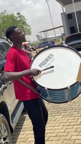 The video captures several men outdoors on a sunny day, engaged in a musical performance. The ground is paved with interlocking bricks, and a modern building with "ARRIVALS" visible on its facade stands in the background, suggesting an airport or similar facility.

In the initial frame (0:00-0:01), a man in a red t-shirt plays a large white bass drum. The drum has "PREMIER" printed on its head, and he holds a drumstick in his right hand. Vehicles are parked behind him, and an airplane can be seen in the distance.

The scene then shifts (0:01-0:02) to three men standing on the paved area. One man wears a green t-shirt with "BEROM BREED" printed on it, another is in a blue polo shirt, and a third is seen from behind in a yellow t-shirt.

Subsequently (0:02-0:04), two men are shown playing trumpets. One wears a white long-sleeved shirt, and the other wears a plaid shirt and a Kufi hat. Both are focused on playing their brass instruments. The bright sunlight indicates it is daytime, and the overall atmosphere appears lively due to the musical activity. The location is in Kwi, Nigeria.