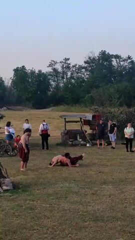 Two men are wrestling in a grassy area while a group of people stands around watching. One man in a red and brown outfit is standing, observing the two wrestlers on the ground. The wrestlers are shirtless and appear to be engaged in a traditional wrestling style, with one man on his knees and the other positioned over him. In the background, there are trees, a wooden structure that looks like a well, and other rustic objects. The sky is bright, suggesting it is either early morning or late afternoon.
