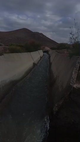 A concrete channel carries water through a dry, hilly landscape under a cloudy sky. The channel is partially filled with flowing water. The surrounding environment consists of dry earth, sparse vegetation, and hills in the background. The lighting suggests it is either dawn or dusk.