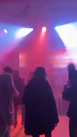 The video shows a concert or party with a large screen displaying "JACQUES" in front of a DJ booth. Several people are in the audience, mostly silhouetted in the dim light. The lighting is a mix of red, white, and blue, creating a party atmosphere. The camera then focuses on a man wearing a black puffer jacket over a yellow hoodie and a blue hat. He throws several small yellow foam objects into the air, appearing to be having fun. The background suggests the event is taking place in a large, open space, possibly a stadium or venue with high ceilings.