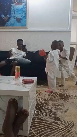 The video shows a living room scene. A man in a white T-shirt and dark shorts sits on a black couch, looking at his phone. Two young boys, dressed in white traditional attire, are playing in front of him. One of the boys holds a red flower bouquet.  A white coffee table with a marble pattern sits in front of the couch, holding a can of soda and a red wallet.  A photo hangs on the wall above the couch. The camera captures a pair of dark-skinned feet at the bottom of the frame. The room is carpeted with a dark and light patterned rug.