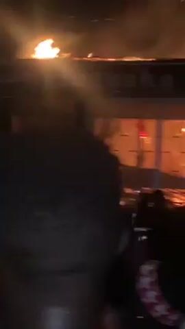 The video depicts a structure engulfed in flames, with a large blaze visible on its roof. Smoke billows into the night sky, and embers are visible in the air. The building, identified by a sign as "CAPITAL PRESSING," is surrounded by several onlookers. The foreground features a blurred dark object, likely the videographer or a nearby object, and silhouettes of individuals can be seen watching the fire. The camera angle is low, looking up at the burning structure. Loud voices are audible in the background.