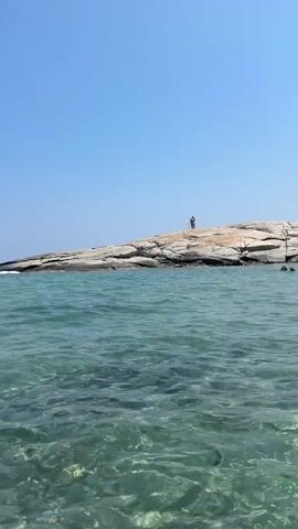 This video showcases a beautiful day at the beach in Castiadas, Italy. The scene is dominated by the crystal-clear, turquoise waters of the Mediterranean Sea, which shimmer under the bright sunlight. The water is shallow enough to see the sandy bottom in places, creating a mesmerizing visual effect.

In the distance, a rocky outcrop juts out from the sea, with two people standing on top, likely enjoying the panoramic view. Further along the coastline, a sandy beach is visible, dotted with lush green vegetation and rolling hills in the background. A few boats, including a sailboat, are out on the water, suggesting a leisurely day for water activities. Several white buoys are strategically placed in the water, possibly marking swimming areas or a course.

The sky is a brilliant, cloudless blue, indicative of a perfect summer day. The overall atmosphere is one of tranquility and relaxation, capturing the essence of a seaside vacation. The time of day appears to be midday, given the strong sunlight and clear sky. There is no visible text in the media. The emotion conveyed is one of peace and enjoyment of the natural beauty of the location.