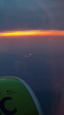 The video shows a view from an airplane window during what appears to be sunset. A portion of the aircraft's green engine nacelle is visible in the foreground. The horizon is illuminated by a band of bright orange and yellow light, indicating the setting sun. Below the horizon, the landscape is dark, with scattered lights visible, suggesting a populated area. The sky above is a deep blue-gray, with some cloud cover. The lighting conditions are low, characteristic of twilight.