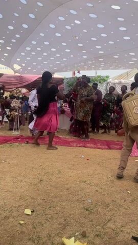 The scene takes place at a wedding or celebration, likely outdoors, under a large white tent with circular cutouts creating a patterned ceiling. Several people are gathered around what appears to be the main area of the event, dancing and celebrating.

A woman wearing a black top and a pink skirt is prominently positioned, moving with her arms raised. Other women are also dancing, dressed in colorful and patterned attire. Some people are standing around observing the festivities.

A young boy with a backpack walks across the foreground of the scene. The ground is covered with a reddish-brown material, possibly dirt or gravel. Red carpets are placed in the center of the area.

In the background, one can see tables, chairs, and floral arrangements, suggesting a reception setup. The overall atmosphere is festive and lively.