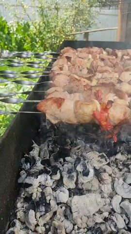 The video shows skewers of meat and tomatoes being grilled over hot coals. The meat is cooking and producing smoke. The scene appears to be outdoors, with greenery visible in the background. The grilling setup includes a metal brazier or grill.