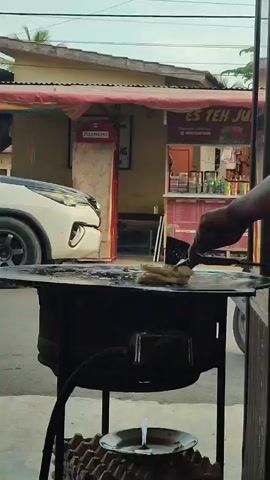 A person's arm and hand are visible on the right side of the frame, using a brush or spatula to cook something on a flat griddle. The griddle is heated by a black stove underneath. In the background, a white car is parked to the left, and a food stall with a red awning is visible. Signs on the stall indicate it sells "ES TEH JUICE" and other beverages. The overall environment appears to be an outdoor setting, possibly a street or market.