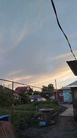 This is a video of a rural landscape in Krasny Gulyai, Russia, during what appears to be dusk or early evening. The sky is filled with soft, wispy clouds tinged with hues of orange and yellow from the setting sun. In the foreground, there is a yard with various objects, including a wooden spool, a blue bucket, and some gardening tools. A garden bed with lush green plants is also visible.

Beyond the yard, several houses are scattered throughout the scene. One house has a red roof, while another is painted a light blue and has a heart symbol drawn on its wall. Trees and other vegetation dot the landscape, adding to the rural atmosphere. A wire runs diagonally across the top right of the frame, suggesting the presence of electrical or communication lines common in such settings.

The overall mood is tranquil and serene, characteristic of a quiet evening in the countryside. There is no discernible human activity taking place in the frame, and no text is visible in the video. The lighting and colors suggest a peaceful transition from day to night.