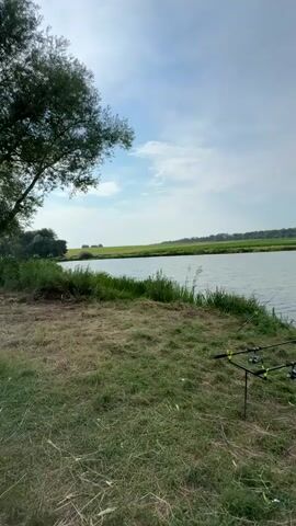 The video shows a scenic view of a body of water, likely a lake or river, with a grassy bank in the foreground. Two fishing rods are set up on a rod pod on the bank, suggesting someone is fishing. The water is calm, reflecting the cloudy sky. In the background, there are trees and green fields. The overall impression is one of a peaceful outdoor setting, likely in the countryside. The camera pans slowly across the scene, capturing the natural beauty of the environment.