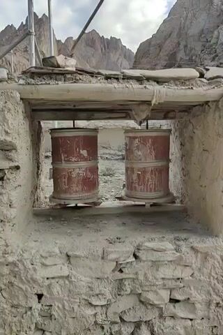 The video shows a close-up view of prayer wheels mounted on the side of a stone building. The prayer wheels are red and weathered, with some signs of rust. They are positioned in an opening in the building's wall, which is made of rough stone and mud. In the background, a dramatic mountain landscape can be seen under a cloudy sky. There are also some scaffolding poles visible on the left side of the frame, suggesting ongoing construction or repair work. The overall impression is one of an ancient religious site in a remote, mountainous region.
