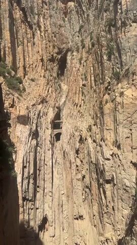 This media file captures a dramatic view of the Caminito del Rey, a famous walkway built along the sides of a narrow gorge in Andalusia, Spain. The primary subject is the immense, towering rock face of the gorge, characterized by its sheer, vertical cliffs and textured, striated surfaces. Sunlight illuminates the rock, casting deep shadows and highlighting the rugged topography.

The scene focuses on a section of the walkway, a narrow path seemingly carved into the cliff face. In the middle ground, a distinctive arched structure is visible, likely a tunnel or a bridge integrated into the path. Vegetation, sparse and hardy, clings to ledges and crevices in the rock, indicating a wild, natural environment. The scale of the cliffs is emphasized by the tiny human-made structures, suggesting a daring feat of engineering in a formidable natural setting.

The activity taking place is implicitly the exploration or traversal of this path. The weather appears clear and sunny, typical of a warm climate, and the time of day seems to be midday, given the bright illumination and sharp shadows. The overall impression is one of awe-inspiring natural beauty and human resilience. There is no visible text in the media file.