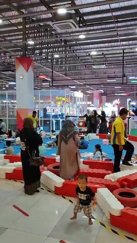 A busy indoor children's play area is depicted. Multiple children and adults are present. In the foreground, a young child in a plaid outfit stands on a tiled floor, facing the camera, holding a small object. The child is barefoot. Behind the child, a low barrier made of red and white plastic segments and tires surrounds a blue mat area. Several adults, some wearing headscarves, are visible near this barrier, some interacting with children. In the background, a yellow-shirted individual appears to be leading an activity with a group of seated children on the blue mat, possibly reading or teaching. Further back, arcade games and playground equipment are visible, along with more people. The environment appears to be a large, open indoor space with a high ceiling, industrial lighting, and visible ventilation systems. The overall atmosphere suggests a public recreational facility for children.