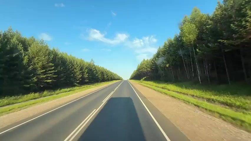 The video shows a forward-facing view of a paved road through a forest. The road has two lanes in each direction, separated by double white lines. Tall, dense evergreen trees line both sides of the road. The sky is blue with scattered white clouds. The sun appears to be shining, casting shadows on the road. The camera is in motion, indicating travel along the road. There are no people or other vehicles visible in the frame. The environment is natural and rural.