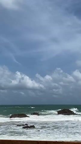 A video captures waves crashing against rocks in the ocean under a partly cloudy sky. The camera is positioned at eye level, looking out towards the sea. In the foreground, a wooden barrier is visible, separating the viewer from the beach. The ocean water is a vibrant turquoise and green, with white foam from the waves swirling around several dark, submerged rocks. The sky is a mix of blue with streaks of white clouds and larger, more ominous gray clouds. The waves are dynamic, with some breaking and sending spray into the air as they hit the rocks. The overall scene is one of natural beauty and the power of the ocean. There are no people or animals visible in the video.
