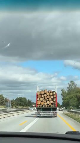The video shows a rear view of a flatbed truck loaded with a large quantity of cut logs. The truck is driving on a highway. The logs are of varying sizes and are stacked high in the truck bed. The truck has a red frame and a license plate that appears to have German characters. The road has yellow lane markings. The sky is partly cloudy with patches of blue sky visible. Trees and vegetation are visible on the sides of the road. Other vehicles can be seen in the distance. The perspective suggests the video is being recorded from another vehicle driving behind the truck.