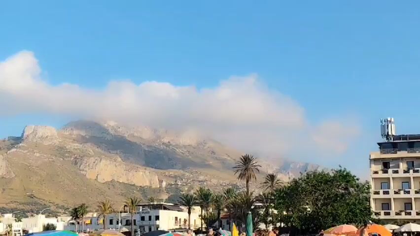 The video pans across a scenic coastal view with a prominent mountain range in the background, partially obscured by dramatic clouds. In the foreground, a bustling beach scene unfolds under a clear blue sky with scattered white clouds. Numerous beach umbrellas in vibrant colors like blue, green, yellow, and orange dot the sandy shore, suggesting a warm, sunny day, likely late afternoon or early evening due to the soft lighting. Several palm trees add to the tropical ambiance.

People are visible on the beach, some under umbrellas, others walking or engaged in beach activities, though their faces are not clearly discernible. A few buildings line the beachfront, including a multi-story hotel with balconies and what appears to be a restaurant or shop with a sign reading "EL REGINA." Another building has a prominent antenna array on its roof. The overall atmosphere is one of leisure and vacation, with the natural beauty of the mountains and the lively beach creating a picturesque setting. The time of day appears to be daytime, with bright sunlight illuminating the scene. The location is identified as San Vito Lo Capo, Italy, a well-known tourist destination characterized by its beautiful beaches and dramatic mountainous backdrop.