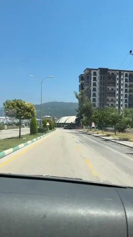 This media file captures a view from inside a vehicle, looking out at a road in Osmaniye, Turkey. The scene is set on a bright, sunny day with a clear blue sky. The road is paved and appears to be a two-lane street with a yellow dividing line. On the left side of the road, there is a well-maintained median strip with manicured green bushes and trees. Beyond the median, more buildings and trees are visible, suggesting an urban or suburban environment.

Dominating the right side of the frame is a tall, modern apartment building with multiple floors and balconies. Trees are scattered along the roadside, casting shadows on the asphalt. There are streetlights lining the road, and several traffic signs are visible, including a yield sign. In the distance, mountains can be seen, providing a scenic backdrop.

The activity depicted is driving, as evidenced by the perspective from inside the car. The road ahead shows some traffic, including what appear to be vans and other vehicles. The overall mood conveyed is one of a typical day in a sunny, possibly warm climate, as suggested by the clear sky and bright lighting. The presence of modern architecture and well-kept landscaping indicates a developed area. No people are clearly discernible as subjects, though there might be individuals in the distant vehicles or within the buildings. The visible text includes that on the traffic signs, which are not legible in detail.