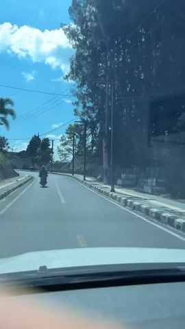 The video shows a view from inside a vehicle driving on a paved road. A motorcyclist is traveling ahead on the same road. The road curves to the left and is lined with a sidewalk. Trees and buildings are visible on the right side of the road. The sky is blue with scattered clouds. Utility poles and wires are also present along the roadside.