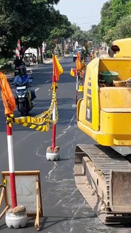 The video shows a street scene with construction activity. In the foreground, a yellow excavator is parked on the right side of the road. Orange and yellow striped caution tape is strung between red and white striped poles, indicating a closed-off area. A small sign on an easel is visible on the left, with some text that is difficult to read but appears to be a warning. The road itself has white markings, possibly indicating construction work or a new surface. In the background, traffic is moving on the road, including cars and a motorcycle. Trees and buildings line the street, suggesting an urban or suburban environment. The sky is clear and the day appears sunny.