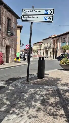 The video shows a street scene in a town with traditional architecture. A street sign indicates parking is available at Plaza del Teatro (150 meters) and Ayuntamiento (400 meters) with arrows pointing to the right. A black trash bin is located near the sign. In the background, several buildings with balconies and tiled roofs line the street. Two cars are parked on the right side of the street, one black and one red. A large circular planter with flowers is positioned on the corner. The sun is shining, casting shadows on the sidewalk. Two people are visible walking down the street in the distance.