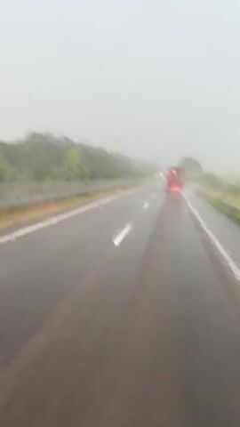A blurry, low-angle, first-person view of a wet highway in what appears to be heavy rain. The road ahead is dark and reflective, with white lane lines visible. Raindrops are visible on the windshield, distorting the view. In the distance, a red truck is seen traveling in the same direction. The surrounding environment consists of green foliage on the sides of the road, obscured by the rain and distance. A windshield wiper is partially visible in the foreground.