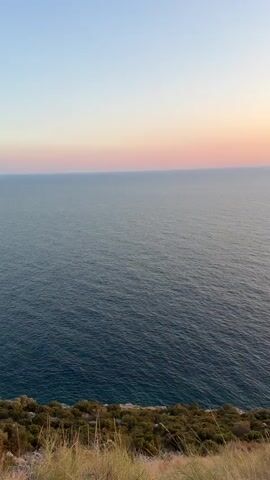 The video shows a wide view of the ocean under a gradient sky of pale blue, pink, and orange, indicating sunset or sunrise. The water is a deep blue with small ripples on the surface. In the foreground, the edge of a grassy cliff is visible, with dry grass and some sparse green vegetation. The camera is static and the view is unobstructed.