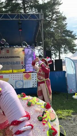 A person dressed as a clown in a red and white striped top hat and a sparkly red and gold costume is on an outdoor stage, blowing bubbles. The clown is facing towards the right of the frame and holding a bubble wand. In the foreground, a large, decorated cake with candy embellishments is visible. In the background, trees and a tent can be seen. The stage has a banner that says "ПРАЗДНИКОМ," which translates to "Happy Holiday" in Russian. The lighting suggests it is daytime, possibly late afternoon due to the angle of the sun.