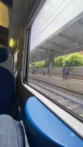Okay, here's a detailed description of the video:

**Setting:** The video is shot from inside a train, looking out of the window at a train platform. The train is stationary at the moment. 

**Environment:** The platform is covered and partially open to the environment. The weather appears bright and sunny. The tracks run parallel to the platform. There's foliage visible just beyond the platform's edge.

**People:** Several people are visible on the platform. Some are sitting, some are standing, and one is walking. There appears to be a group of people sitting together, possibly waiting.

**Objects:** Various elements are visible on the platform, including signage, metal structures, and luggage. There's a digital information display showing train times and destinations. Inside the train, a blue seat and the jeans-clad leg of a passenger are visible.

**Actions:** The train is currently stopped at the station. People on the platform are either waiting or passing through.

**Overall Impression:** The scene captures a typical moment at a train station on a bright day, with passengers going about their business while waiting for or after arriving on the train.