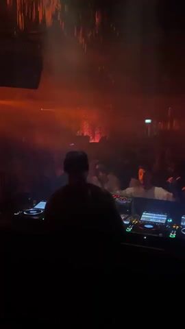 A DJ is shown at a console with multiple decks and screens. The DJ is wearing a dark baseball cap and a dark shirt. The DJ is facing away from the camera, towards a crowd of people. The crowd is dancing and enjoying the music in a dimly lit club. There are red and yellow lights illuminating the scene. Some people in the crowd are visible in the background, and some are blurry due to motion. The overall atmosphere appears to be energetic and celebratory.
