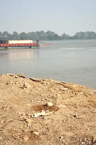 The video shows a wide river with a barge carrying two shipping containers and some lighter colored cargo. The barge is being pushed by a boat. The shore in the foreground is sandy with some rocks and vegetation. The background features trees and a hazy sky. Another smaller boat is visible on the right side of the river.