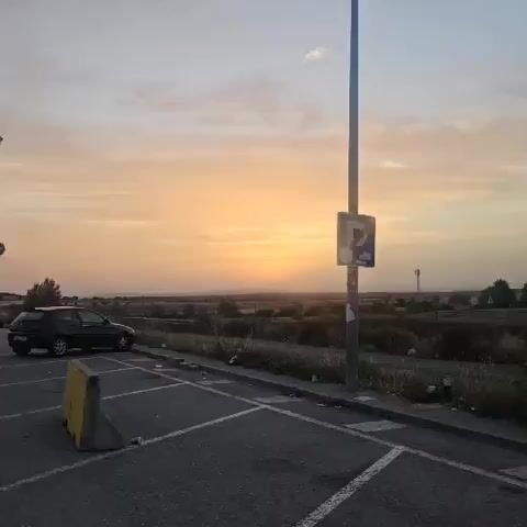 The video shows a sunset over a rural landscape. The sun is low on the horizon, casting a warm, orange glow across the sky. In the foreground, there is a road and a lamppost with a parking sign. The landscape is dotted with trees and bushes. The sky is mostly clear, with a few wispy clouds. The overall impression is one of peace and tranquility.