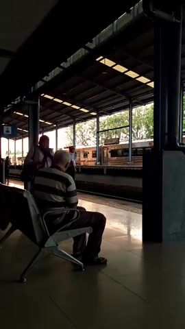 Here is a description of the video:
The video shows a train station platform. An older man is sitting on a metal chair, facing away from the camera. A woman in a headscarf is standing next to him, adjusting something on his back. Behind them, a train is visible on the tracks, and other people are walking around the platform. The platform has a tiled floor and a roof supported by metal pillars. The lighting is dim, but natural light is coming in from the open side of the platform.