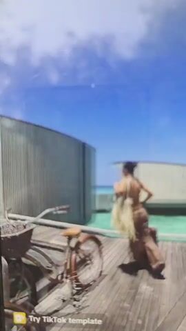 A woman in a bikini and matching pants walks along a wooden dock towards a building. She enters the building and walks through a hallway into a bedroom. The room has a bed, a dresser, and a window with a view of the ocean. The woman walks around the room.