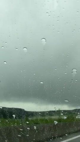 The video shows a view through a car windshield on a rainy day. Raindrops are visible on the glass, obscuring the view to some extent. The background consists of a highway with green grass and trees in the distance. A white van and another vehicle with flashing red and blue lights can be seen driving on the highway. Streetlights are also visible along the roadside. The sky is overcast and gray.