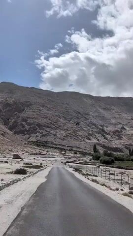 The video shows a paved road traveling through a desert landscape. The road is flanked by barren, sandy terrain with sparse vegetation and a chain-link fence. In the background, rugged, rocky mountains rise under a cloudy sky. The camera appears to be moving along the road, capturing the expansive, arid environment.