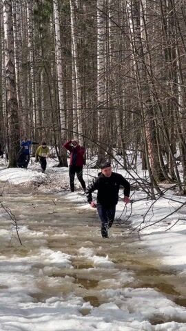 The video captures a scene in a snowy, wooded area where a group of people are participating in some sort of race or event. The environment is characterized by snow-covered ground, scattered ice patches, and a thin stream of water flowing through the area. Several bare trees stand tall in the background.

In the foreground, a man wearing a black hooded sweatshirt and dark pants runs through the stream of water, smiling as he looks towards the camera. Behind him, three other people are visible; one wearing a bright yellow shirt and another in blue. The person in the red jacket appears to be holding a camera, possibly documenting the event.

The individuals, who seem to be participants or spectators, are interacting with the natural environment. The focus is on the person running through the water, exhibiting an expression of enjoyment as he tackles the icy obstacle.