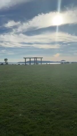 The video opens with a wide shot of a grassy field under a partly cloudy sky with the sun shining brightly. In the distance, across a body of water, a treed shoreline is visible. A large, modern structure with large pillars supporting a horizontal beam stands in the mid-ground. To the right of this structure, there are people gathered under a canopy. The camera pans slowly to the right, revealing more of the grassy field and the people under the canopy, and then pans back to the left. A person is seen walking in the field. The sun is high in the sky, creating a lens flare effect in some shots. The overall atmosphere is serene and appears to be a public park or waterfront area.