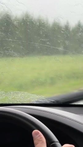 The video shows a view from inside a car through a windshield. The windshield is wet with rain, and the windshield wipers are moving to clear the view. A person's hand is visible on the steering wheel. Outside the car, there is a green field and trees in the background. The sky appears overcast. There is a small electronic device mounted on the dashboard. The overall impression is that the car is driving in rainy weather.