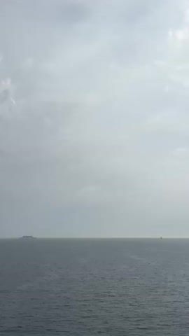 The video shows a view of the ocean under a cloudy sky. The sea is gray and calm. The sky is mostly overcast with varying shades of gray clouds. The sun is visible, shining brightly through the clouds.
