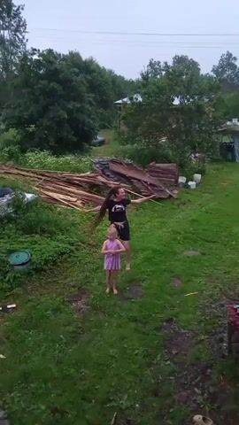 Two people, one adult and one child, are dancing in a grassy yard. The adult is wearing a black t-shirt and dark shorts. The child is wearing a purple dress. There are trees and buildings in the background. There is also a pile of lumber in the yard.