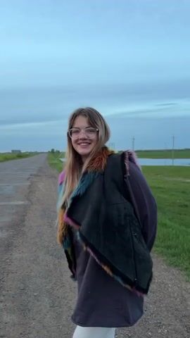The video shows a young woman with long blond hair and glasses, standing on a dirt road next to a flooded green field. She is wearing a long-sleeved shirt, leggings, and a dark, fur-trimmed vest. The field is largely covered with water and dotted with power poles. The sky is overcast. She smiles and laughs before the camera pans right to display the flooded landscape.