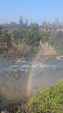 The video shows a wide waterfall on a sunny day. A rainbow is visible at the base of the falls near some greenery. The water flows rapidly over rocks and forms mist at the bottom. Trees and buildings are visible in the background. The sky is blue with some clouds.