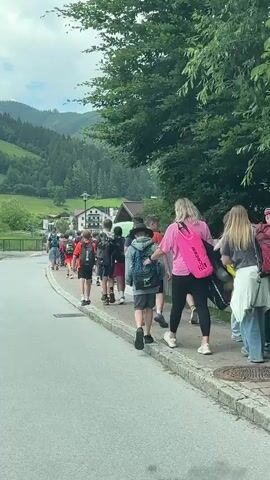 A group of approximately fifteen people, mostly children and adolescents, are walking away from the camera on a paved sidewalk next to a road. They are wearing casual clothing and many are carrying backpacks. They are walking in a line, and appear to be moving towards the left of the frame. The environment is a rural mountainous area with green hills covered in trees and some scattered buildings. The sky is cloudy.